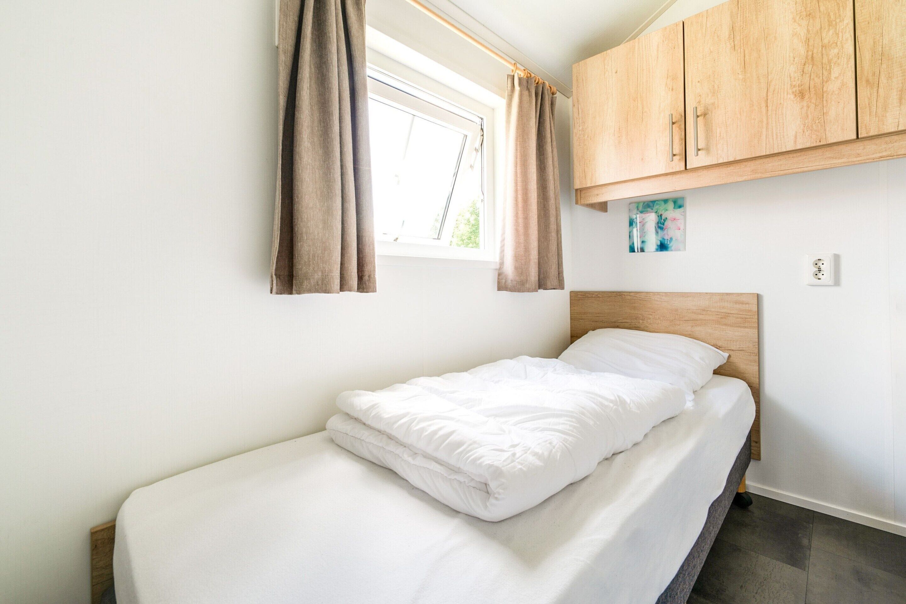 A single bed with white bedding in a room with a wooden headboard, a window with curtains, and a wooden cabinet above the bed.