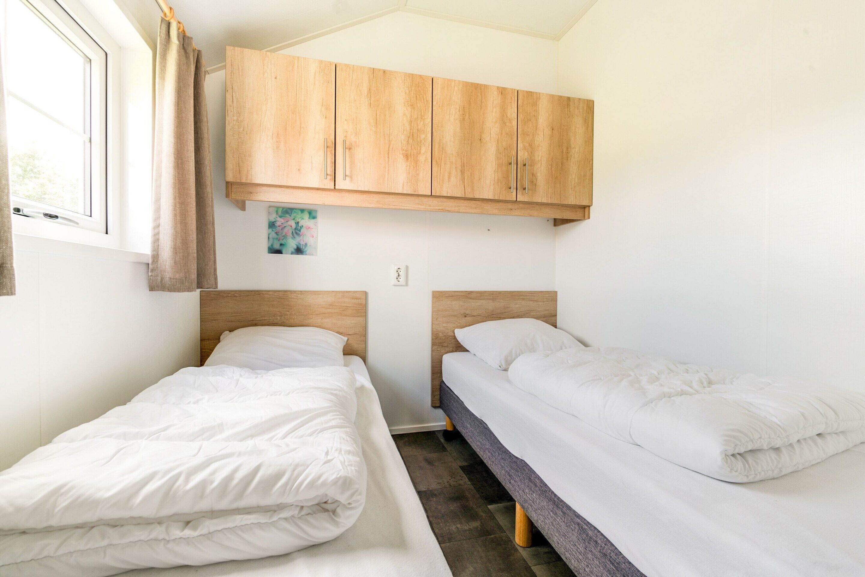 Une petite chambre propre, avec deux lits simples, des armoires en bois au-dessus et une fenêtre avec des rideaux.