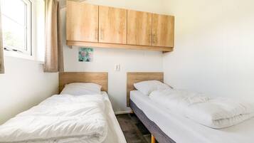 Une petite chambre propre, avec deux lits simples, des armoires en bois au-dessus et une fenêtre avec des rideaux.