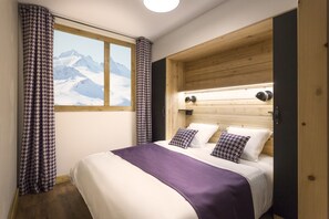 A bedroom with a wooden headboard, a large bed with white and purple bedding, a view of mountains through a window, and houndstooth curtains. - Mountain Stay With Spa (Tignes)