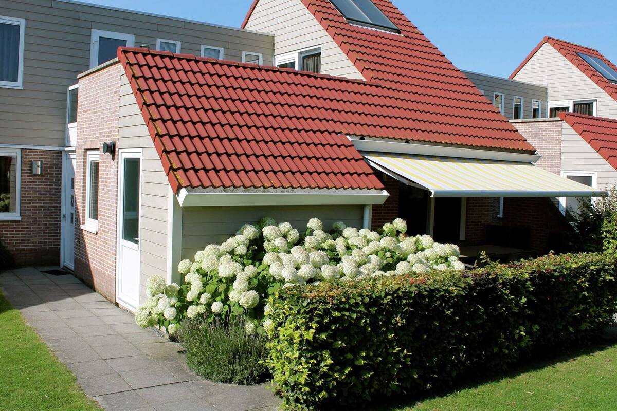 Ein zweistöckiges Haus mit rotem Ziegeldach, einer kleinen angebauten Garage und einer Veranda mit weißen Blumen.