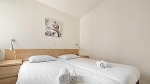 A neatly made bed with white linens and rolled towels, a wooden headboard, a bedside table with a lamp, and a wall-mounted artwork. - Well-kept Bungalow With Microwave Near Valkenburg (Walem)