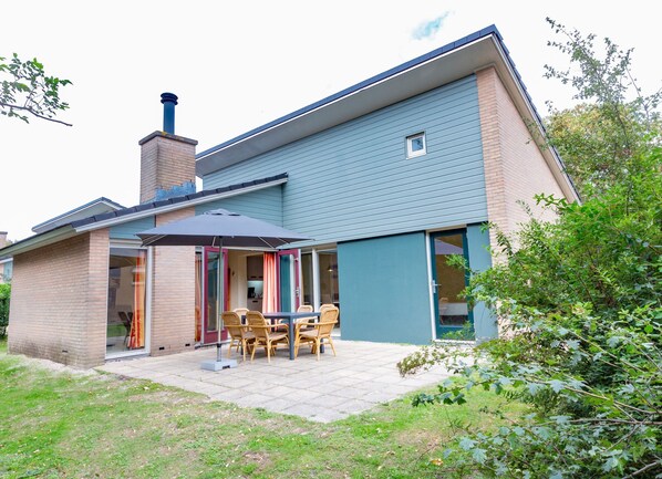 A two-story house with a patio area featuring a table and chairs, a chimney, and a small window. - Restyled Holiday Home with Fireplace near Beach (The Hague)