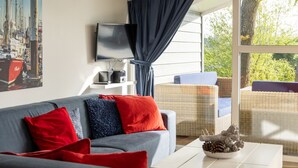 A modern living room with a grey sofa, red and blue cushions, a flat-screen TV, a coffee table with a pinecone decoration, and a wicker chair. - Modern Chalet Near Lake Sneek (Terkaple)