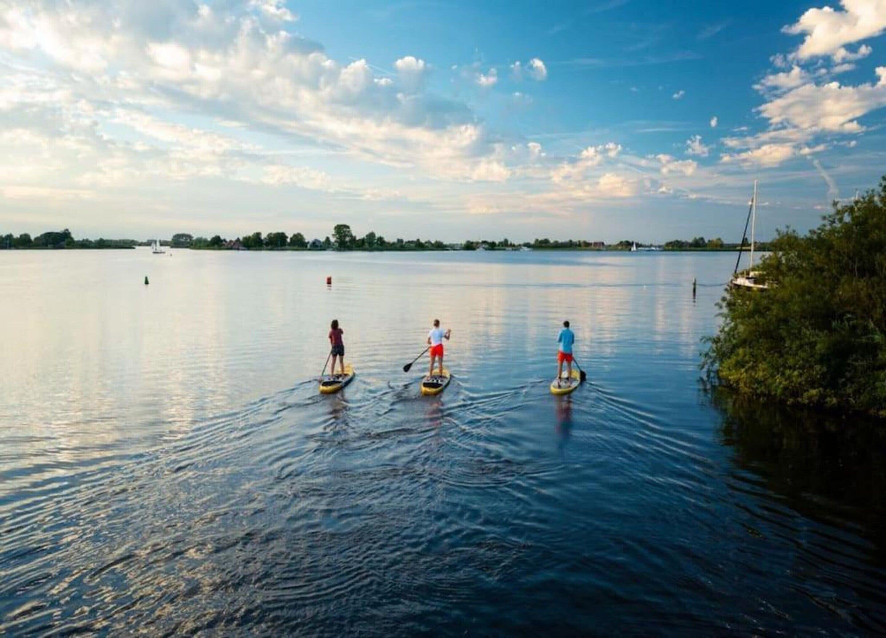 Três pessoas praticando paddleboard em um lago calmo, com céu claro e barcos ao longe.