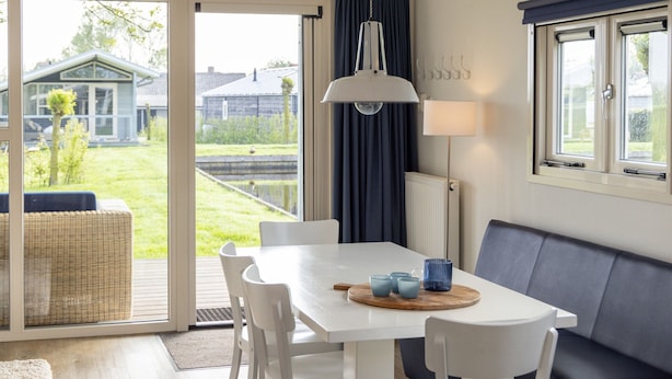 A modern dining area with a white table, blue chairs, a wooden board with cups, a hanging lamp, and a view of a house through a glass door. - Modern Chalet Near Lake Sneek (Terkaple)