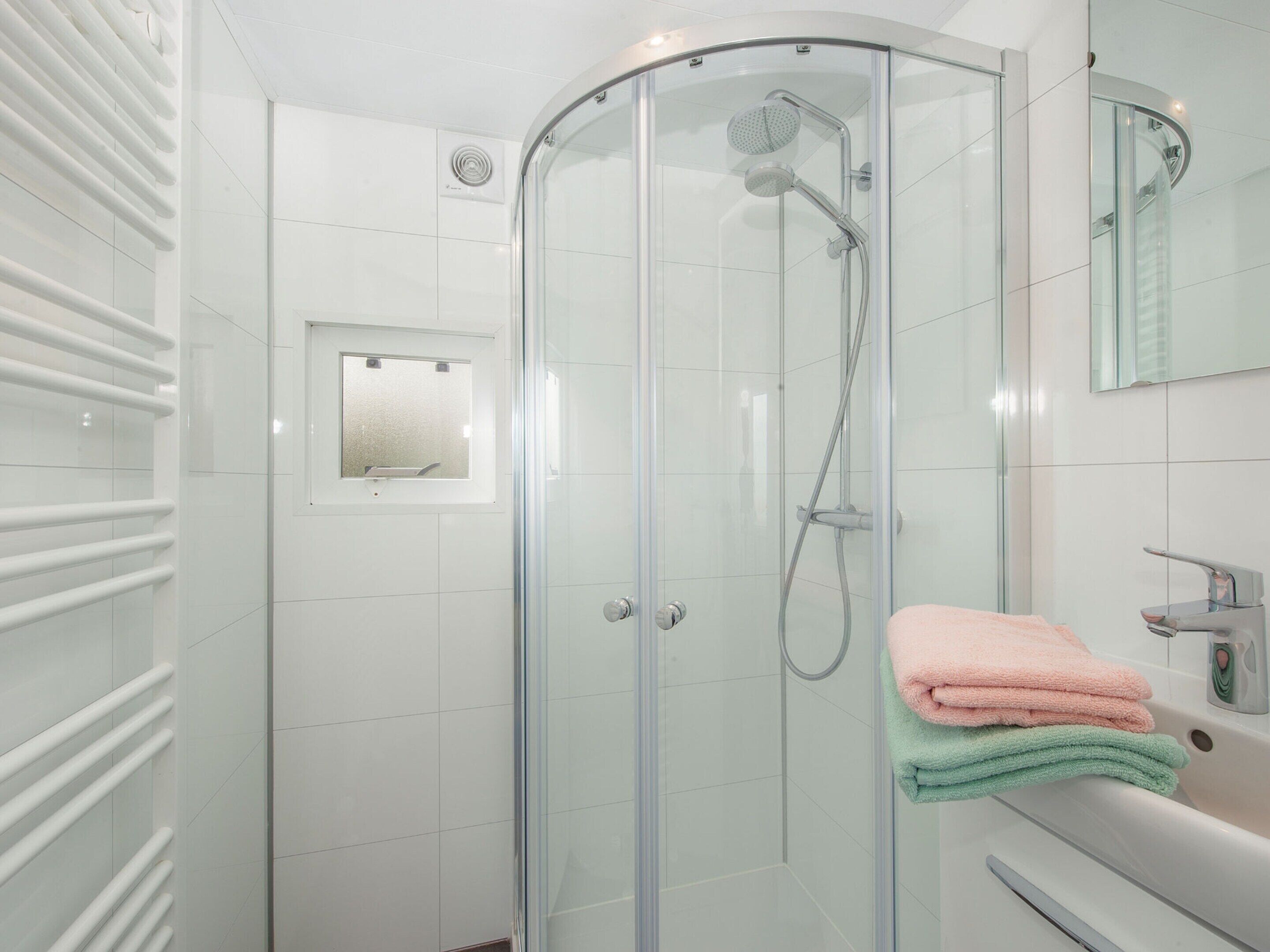 A modern bathroom with a glass-enclosed shower, a white tiled wall, a small window, and a sink with a mirror.