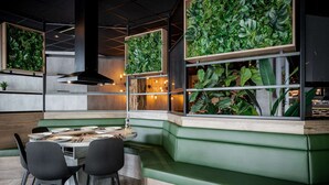 Un intérieur de restaurant moderne avec un coin salon composé de banquettes vertes, des tables en bois et de grandes fenêtres ornées de murs végétaux.