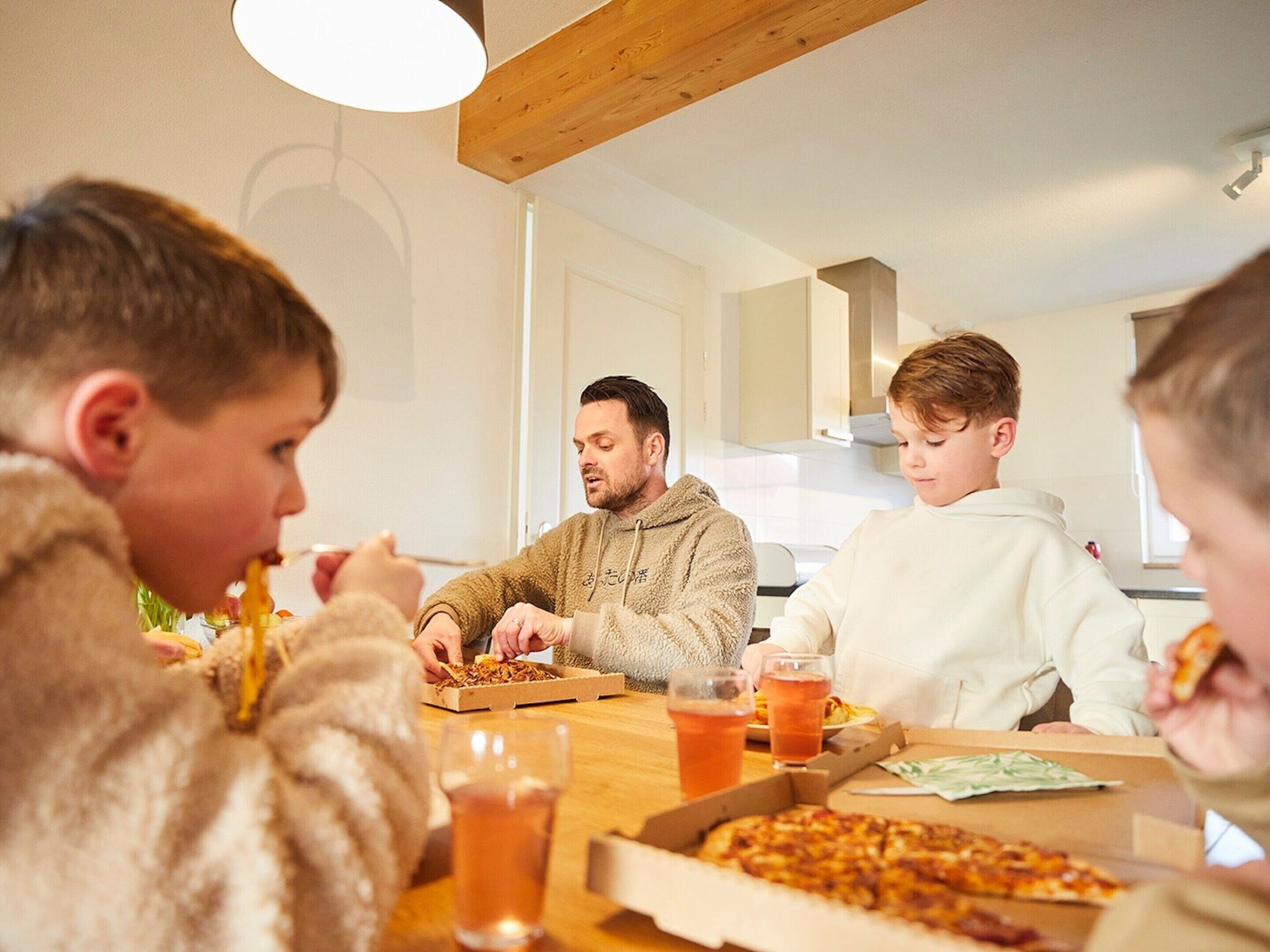 Een gezin zit samen aan tafel met pizza dozen en drankjes.
