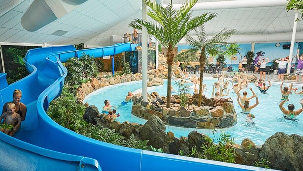 Un parc aquatique intérieur doté d’un toboggan bleu, d’un espace piscine entouré de rochers et de palmiers, et où les visiteurs profitent des installations.