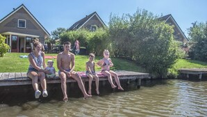 A group of people sitting on a wooden dock by a calm body of water, with houses in the background. - Chic Stay by the Water (Medemblik)