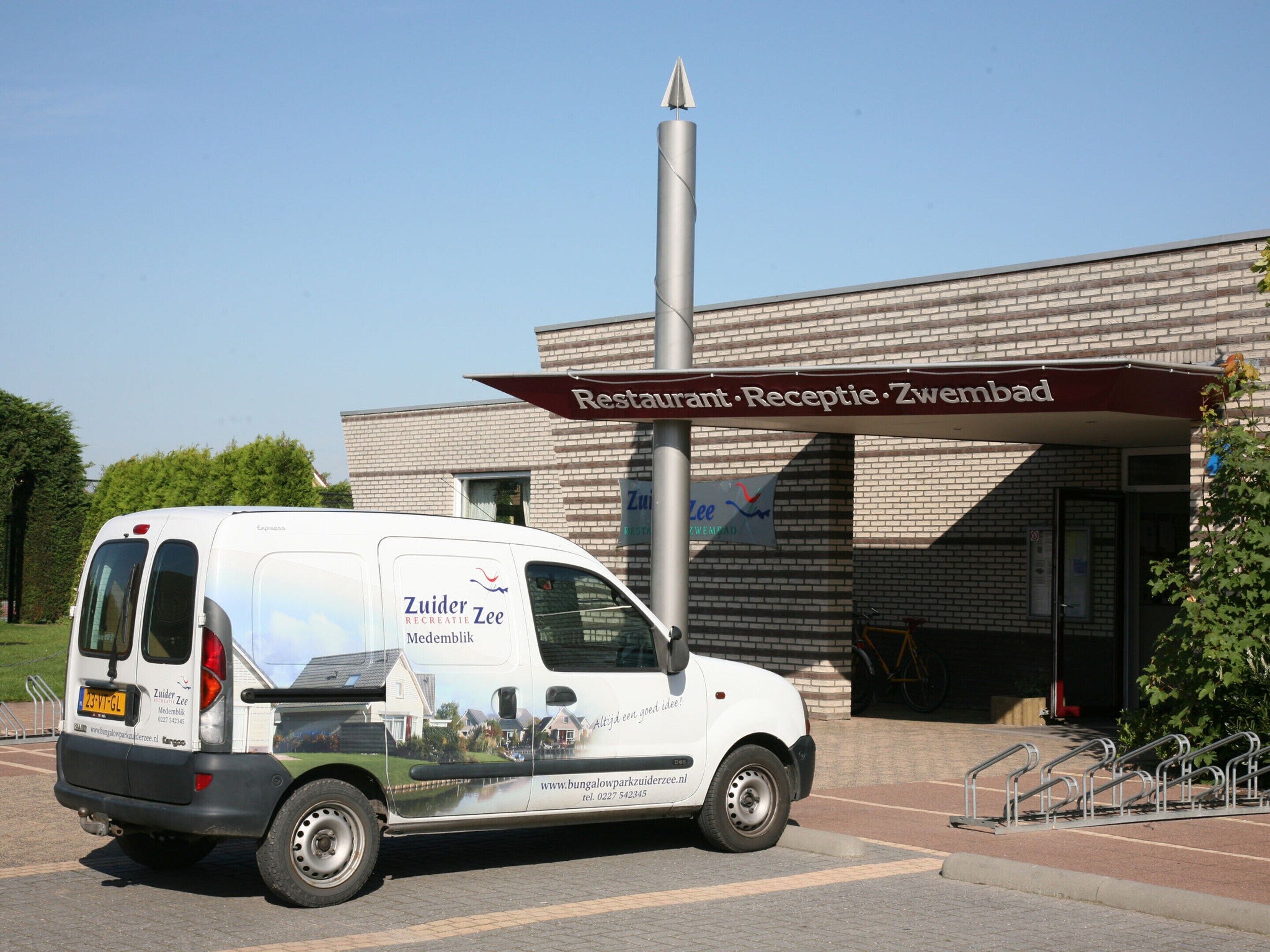 Een witte bestelwagen geparkeerd voor een gebouw met een bord waarop staat: 'Restaurant, Receptie, Zwembad'.