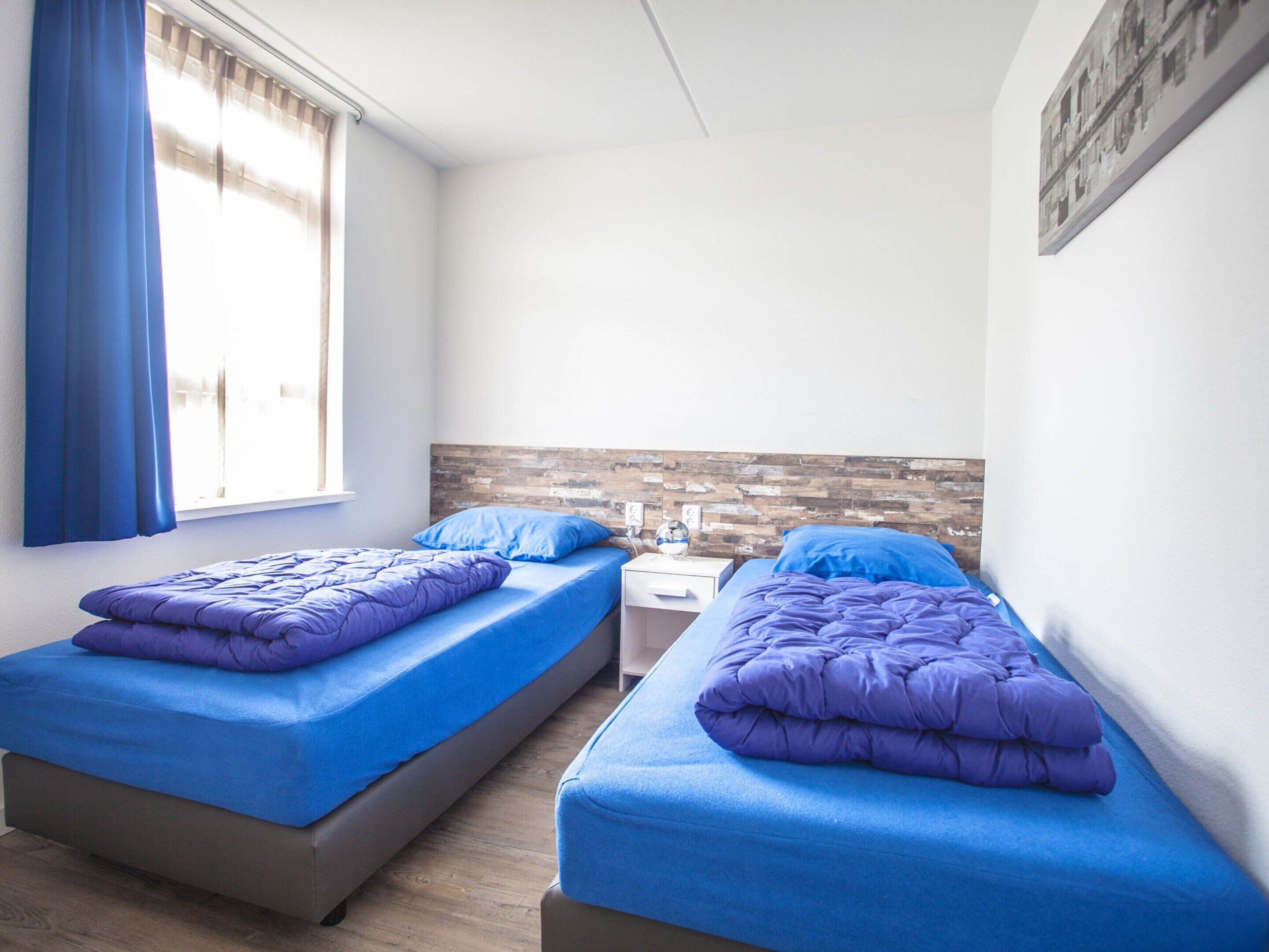 A room with two beds, each with a blue mattress and purple bedding. There is a window with blue curtains, a wooden headboard, and a small table with a lamp.