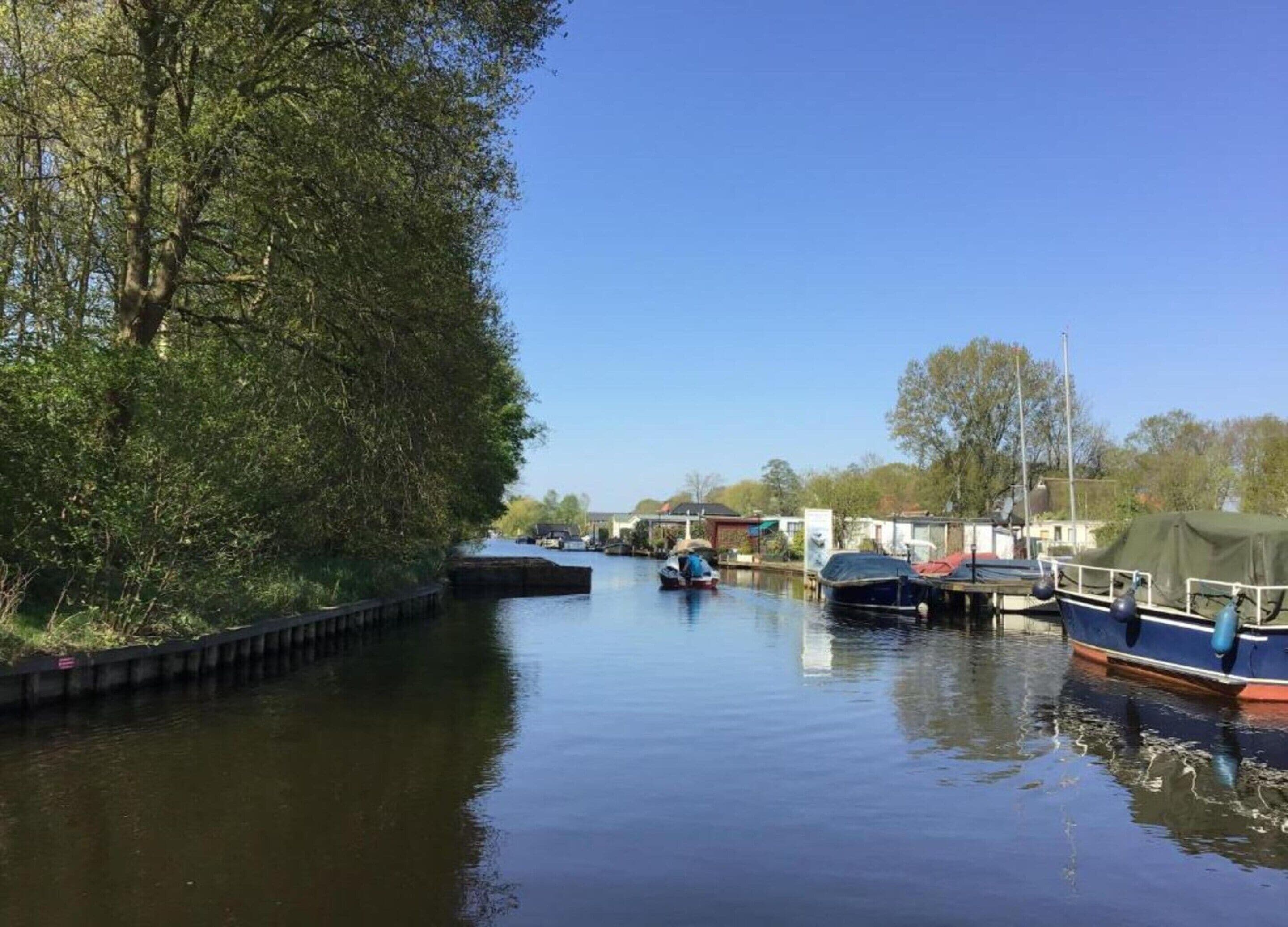 A calm canal with boats docked on the right, surrounded by trees and buildings in the background.