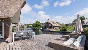 Eine Holzterrasse mit Gartenmöbeln, ein Gebäude mit Strohdach und Blick auf einen Kanal.