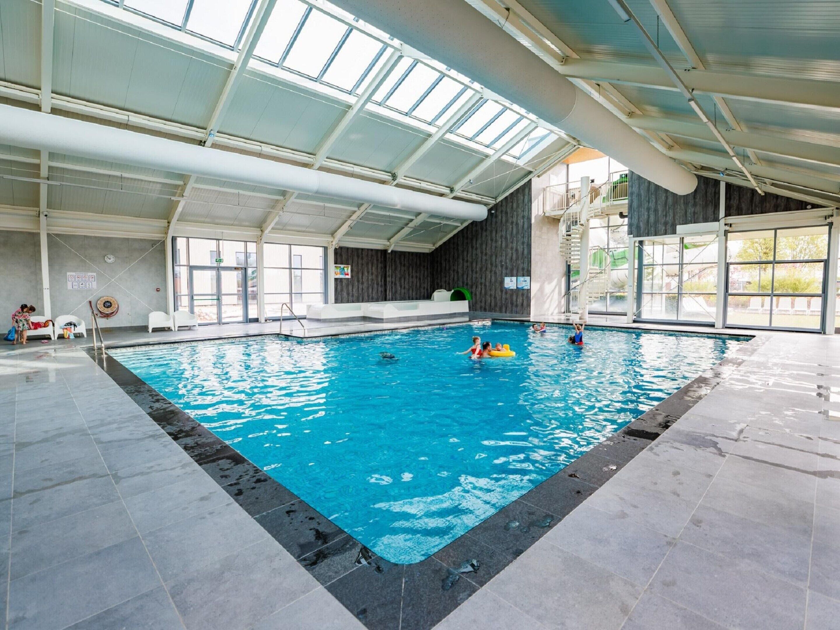 Una piscina interior con agua cristalina azul, suelo de baldosas y un techo alto con tragaluces.