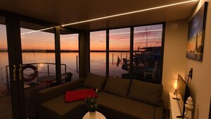 Ein modernes Wohnzimmer mit einem großen Fenster mit Blick auf eine Marina bei Sonnenuntergang.