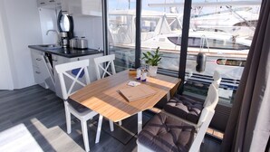 Eine moderne Küche mit weißen Schränken und einer Kaffeemaschine, ein Holzesstisch mit einem Notizbuch und Blick auf Boote durch große Fenster.