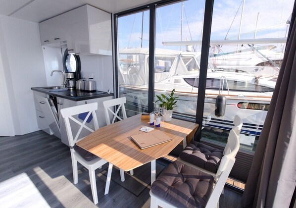 Una cocina moderna con armarios blancos y una cafetera, una mesa de comedor de madera con un cuaderno, y vistas a barcos a través de grandes ventanales.
