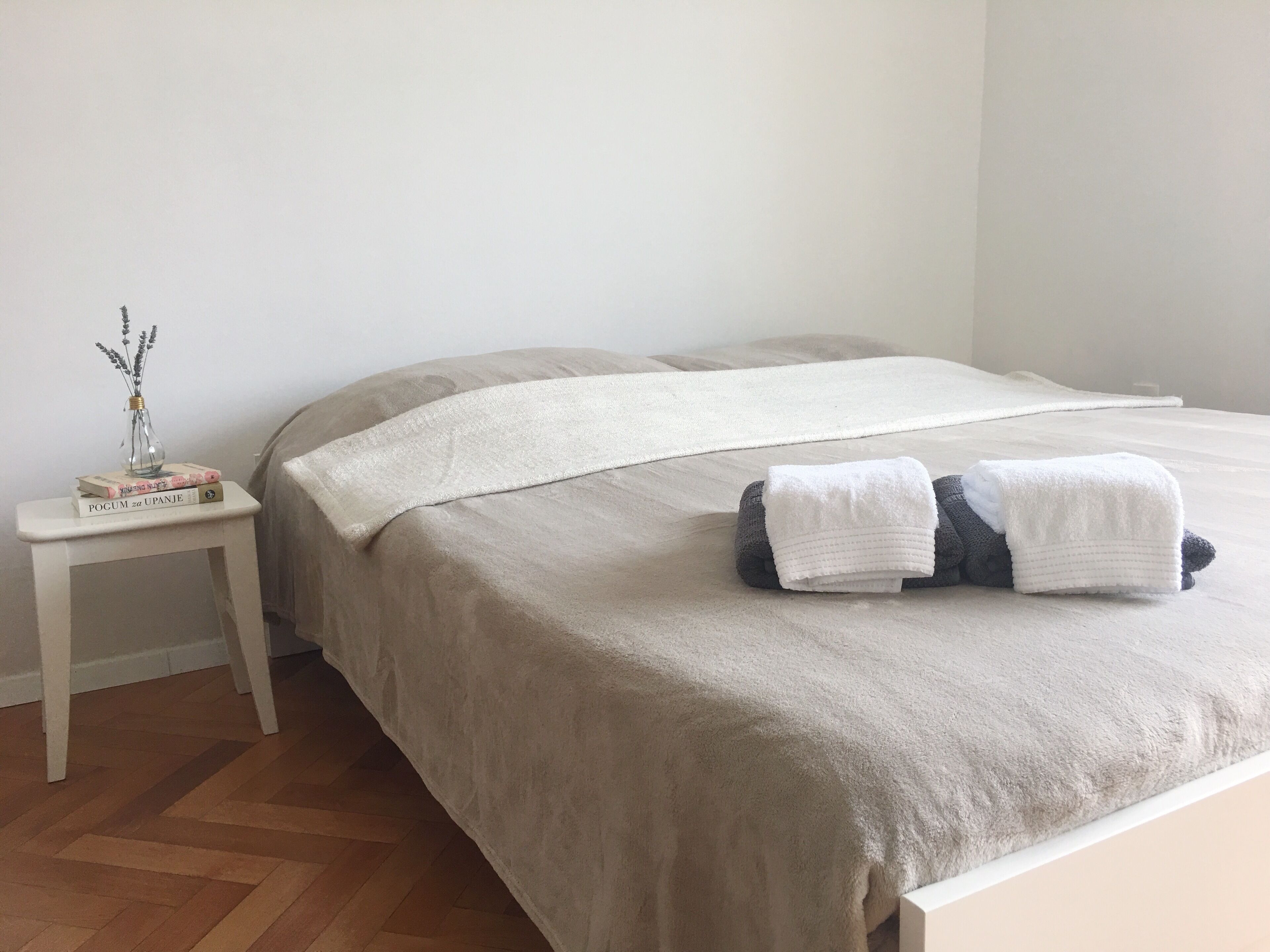 A neatly made bed with a beige blanket and pillows, a bedside table with a vase and books, and a wooden floor.