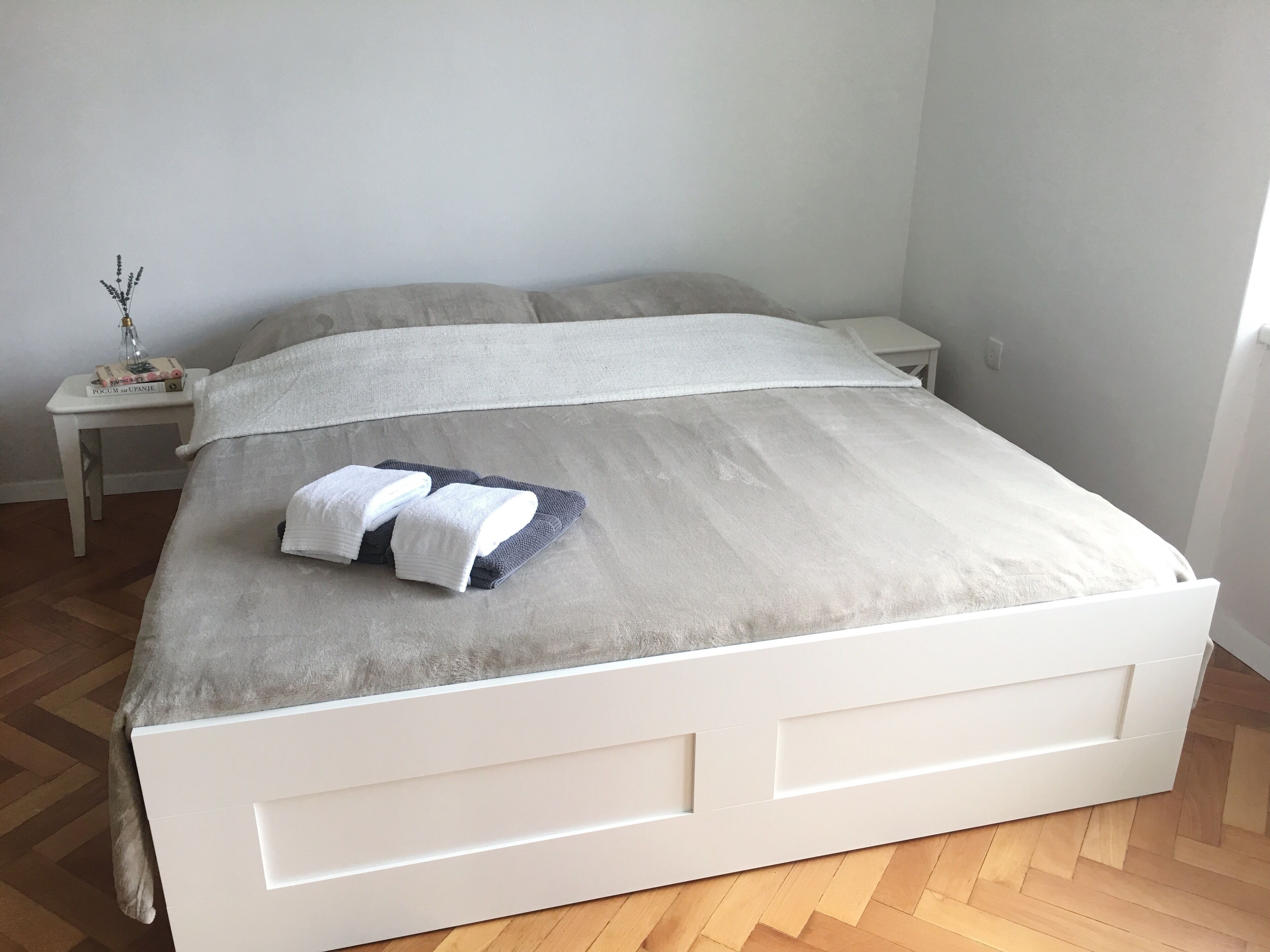 A neatly made bed with a grey duvet, white pillows, and folded towels on a white headboard. There is a small bedside table with a book and a decorative item. The room has wooden flooring and light-colored walls.