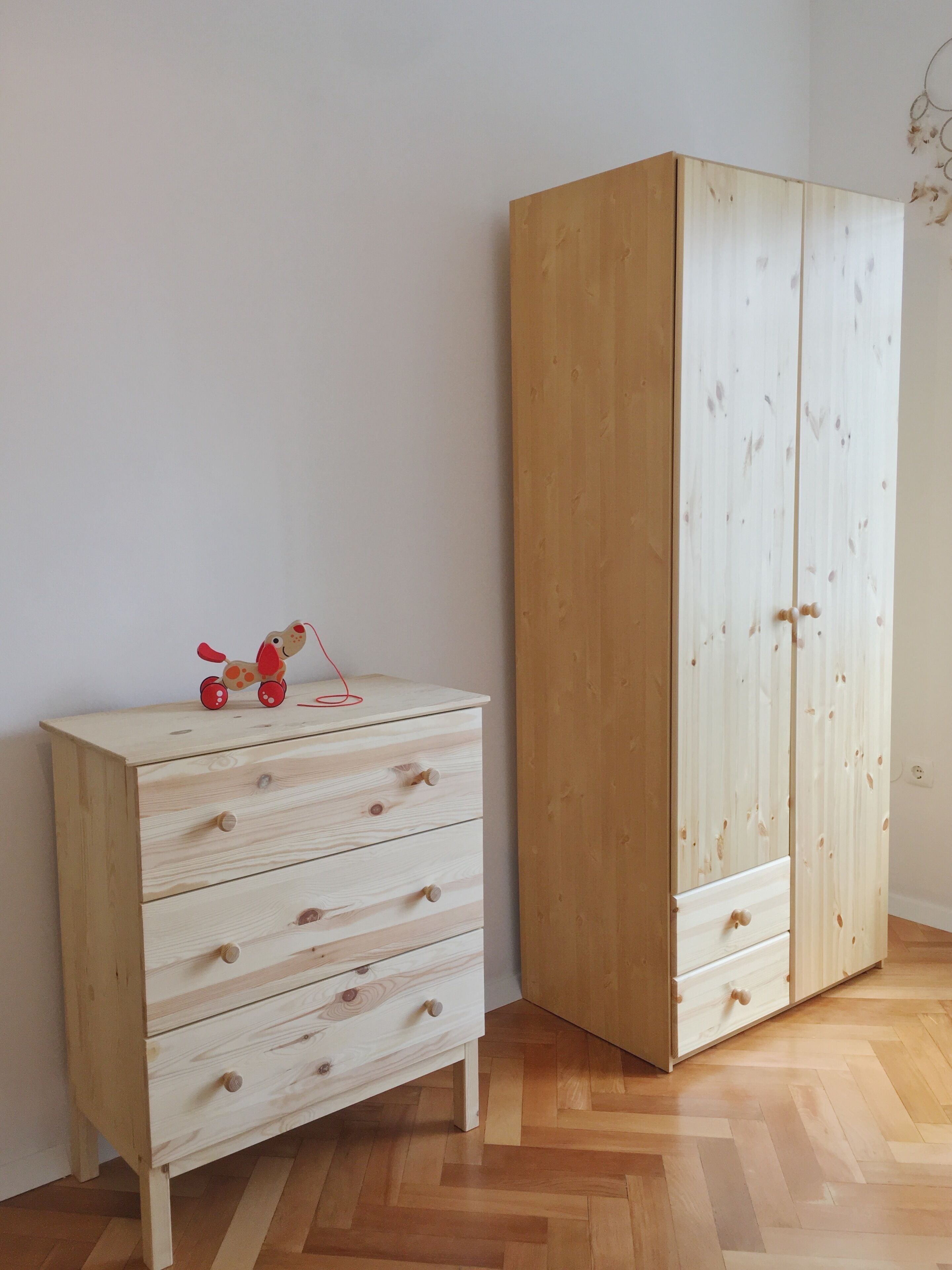 Ein Zimmer mit Holzboden, einer kleinen weißen Kommode mit Schubladen und einem hohen Kleiderschrank mit zwei Türen.