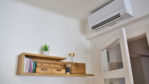 A room with a white wall, a wooden shelf with books and a small plant, and a white air conditioning unit mounted on the wall.