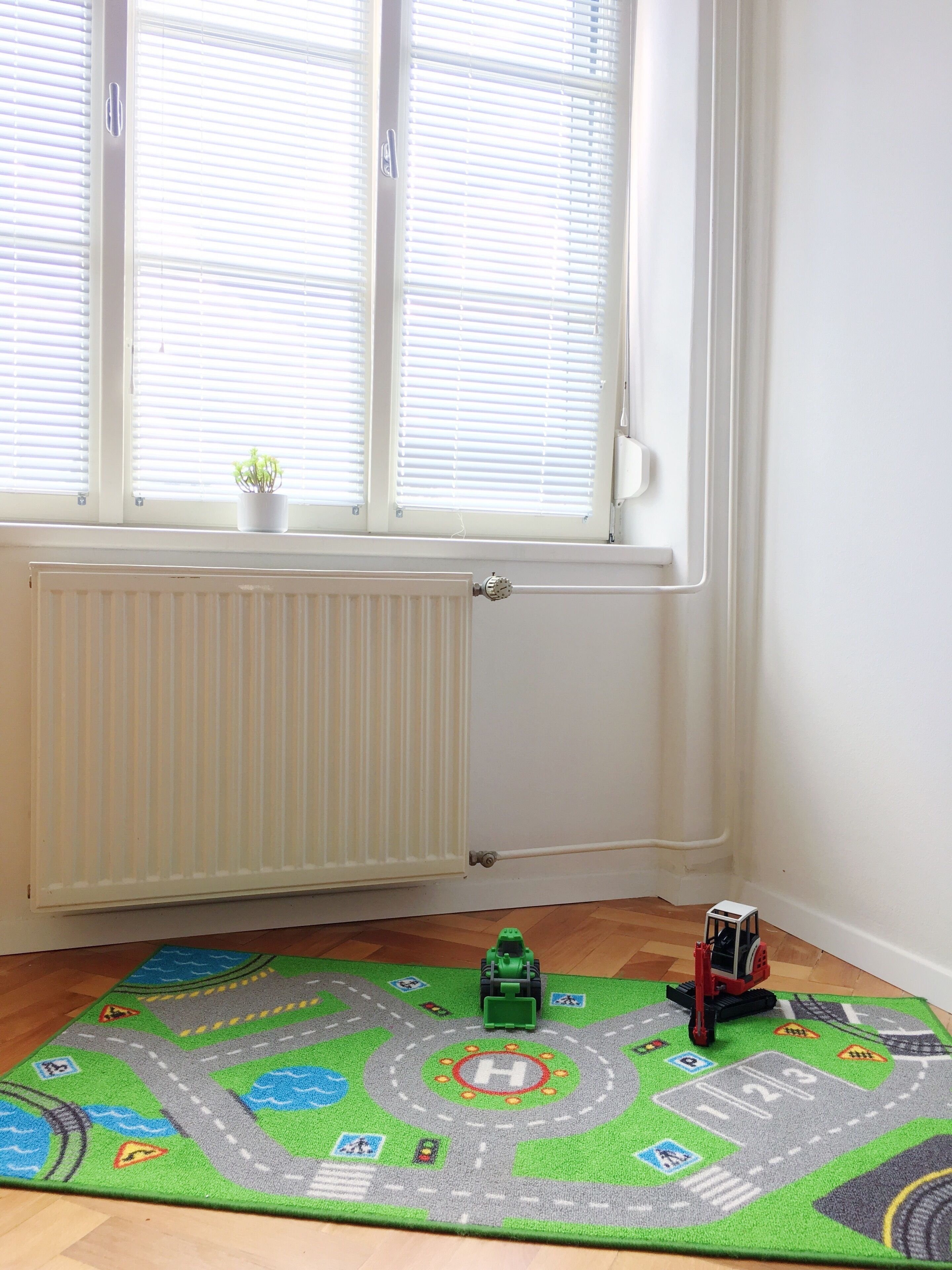 Una habitación con ventana, radiador y un tapete de juego con símbolos de carreteras y vehículos.
