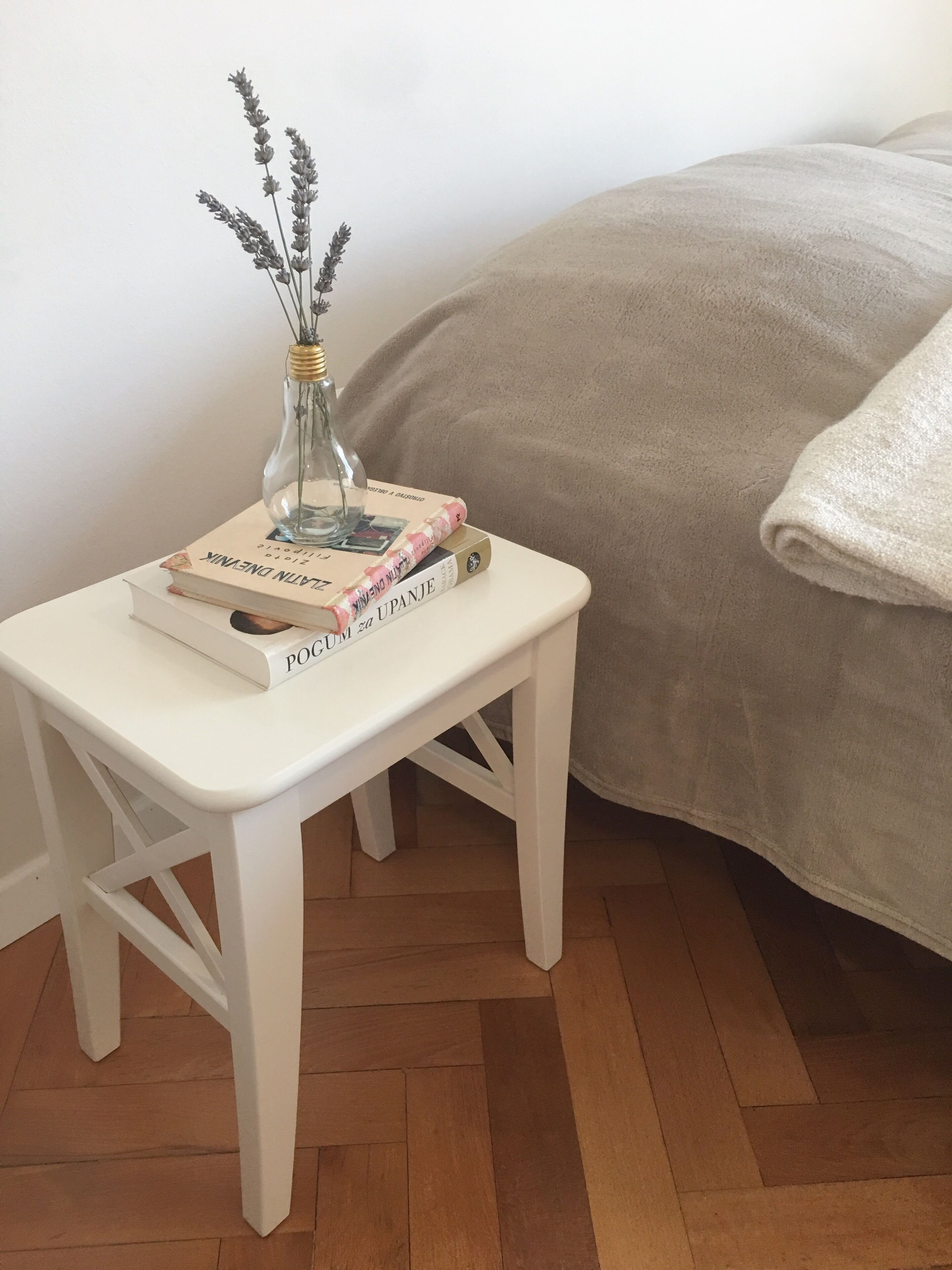 A white bedside table with a glass vase containing lavender, a stack of books, and a folded towel.