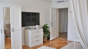 A modern bedroom with a flat-screen TV, a white dresser, a potted plant, and a large window.