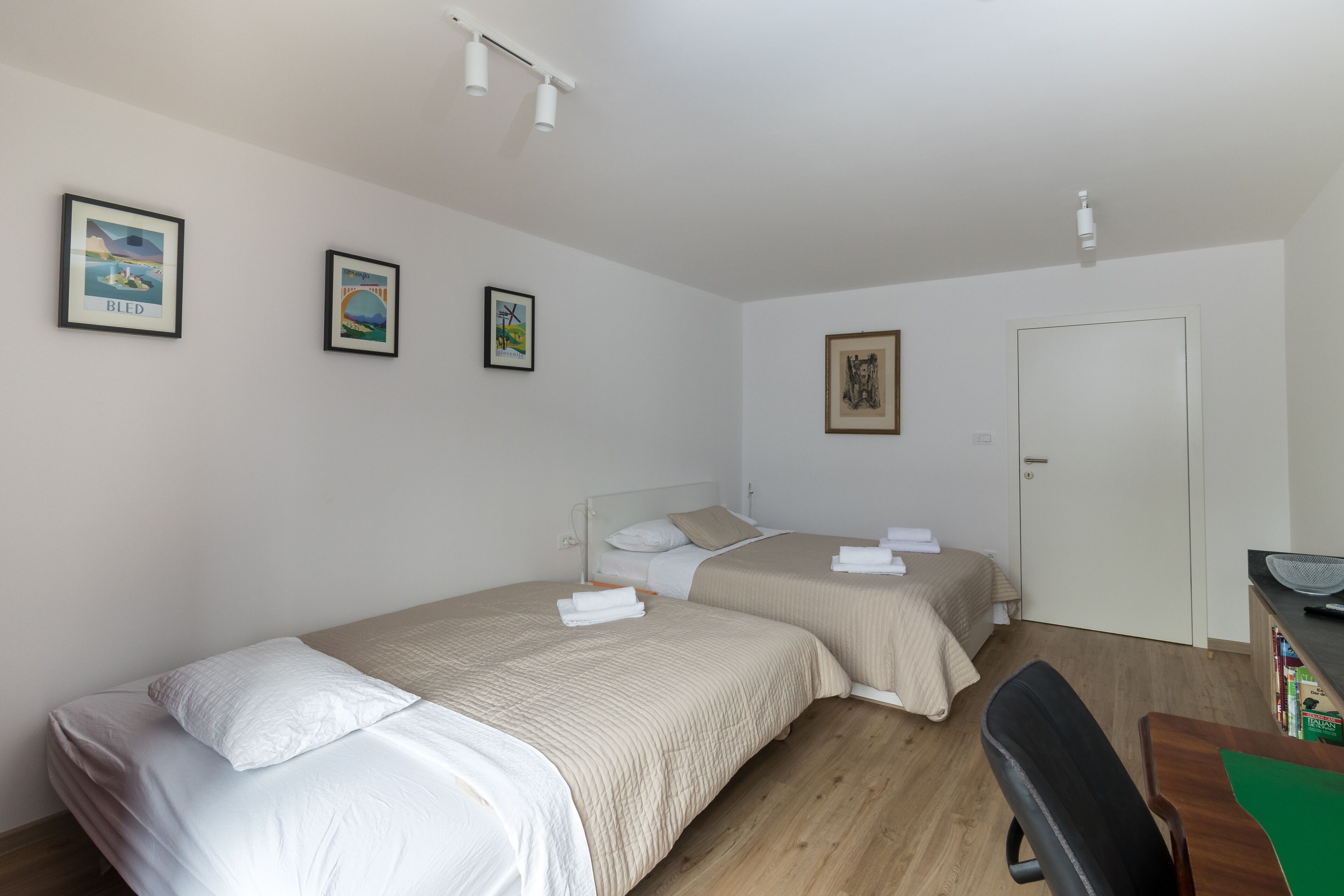 A room with two beds, a desk, and a door. There are framed pictures on the wall.