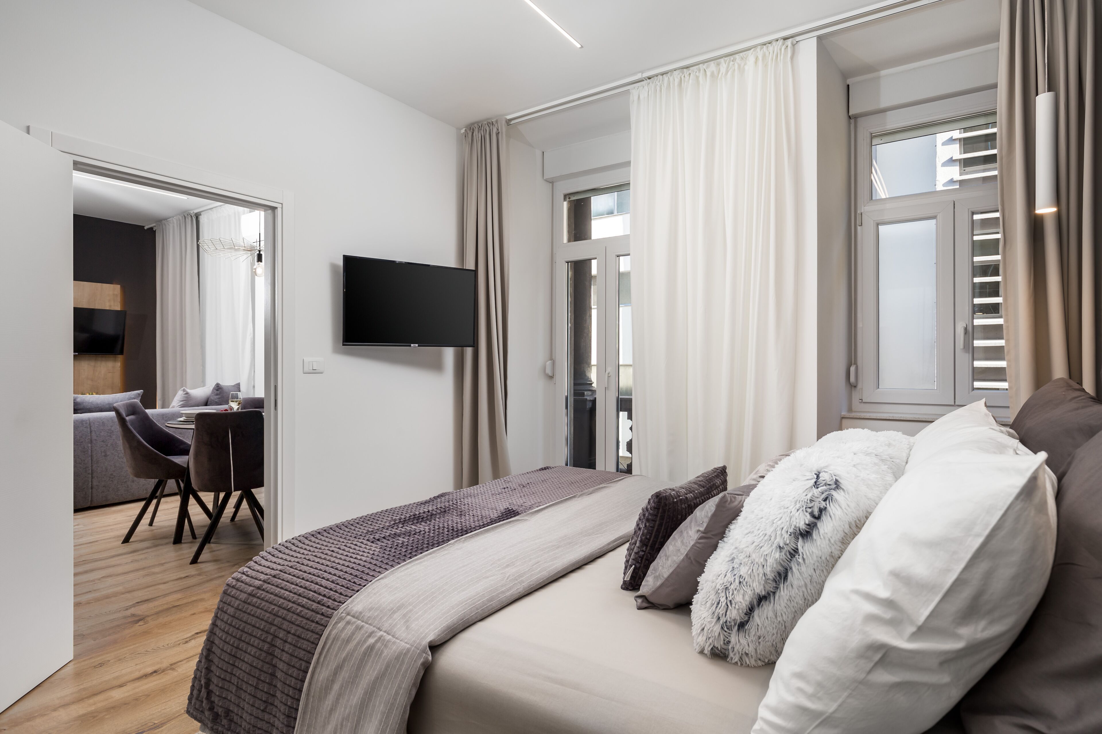A modern hotel room with a large bed, a flat-screen TV, a dining area with a table and chairs, and a view of the city through the window.