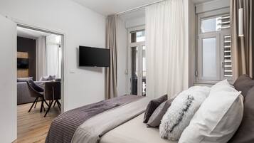 A modern hotel room with a large bed, a flat-screen TV, a dining area with a table and chairs, and a view of the city through the window.