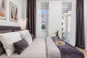 Un dormitorio con una cama, almohadas, una mesita de noche con un florero y un plato de croissants, y vistas de edificios a través de una ventana.