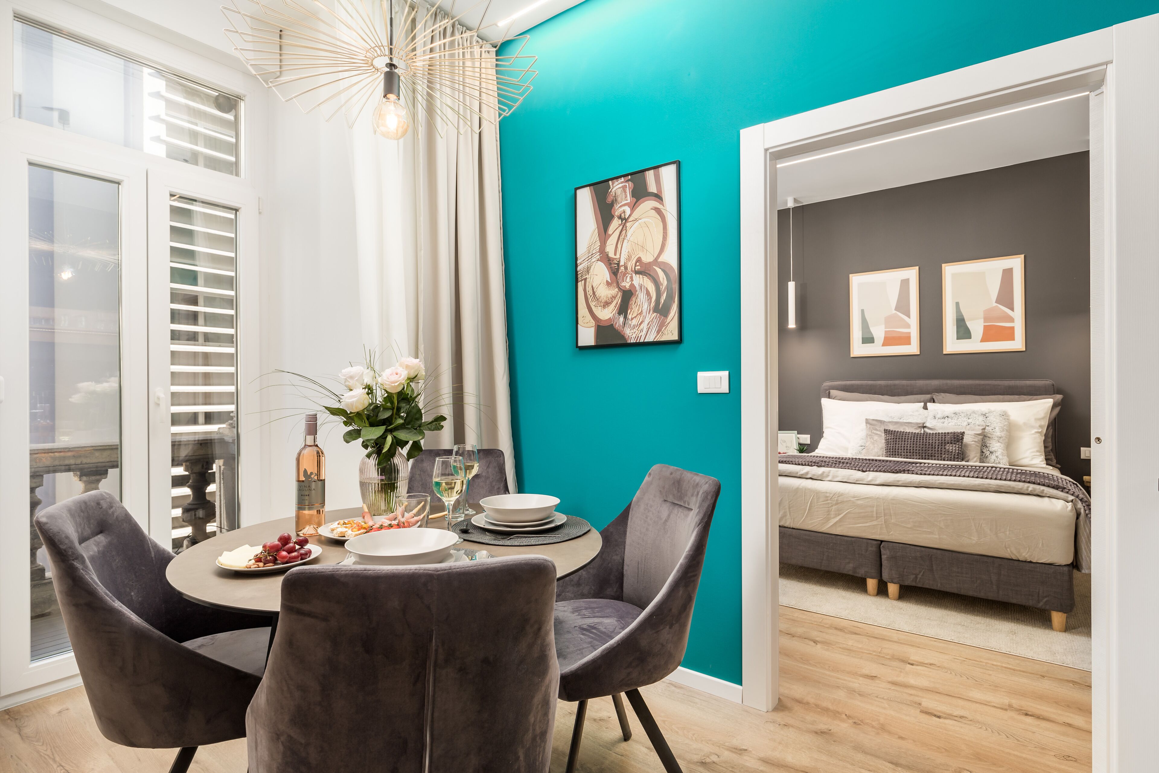 Een moderne woonkamer met een turquoise accentmuur, een eethoek met een ronde tafel en stoelen, en een slaapkamer met een bed en kunstwerken.