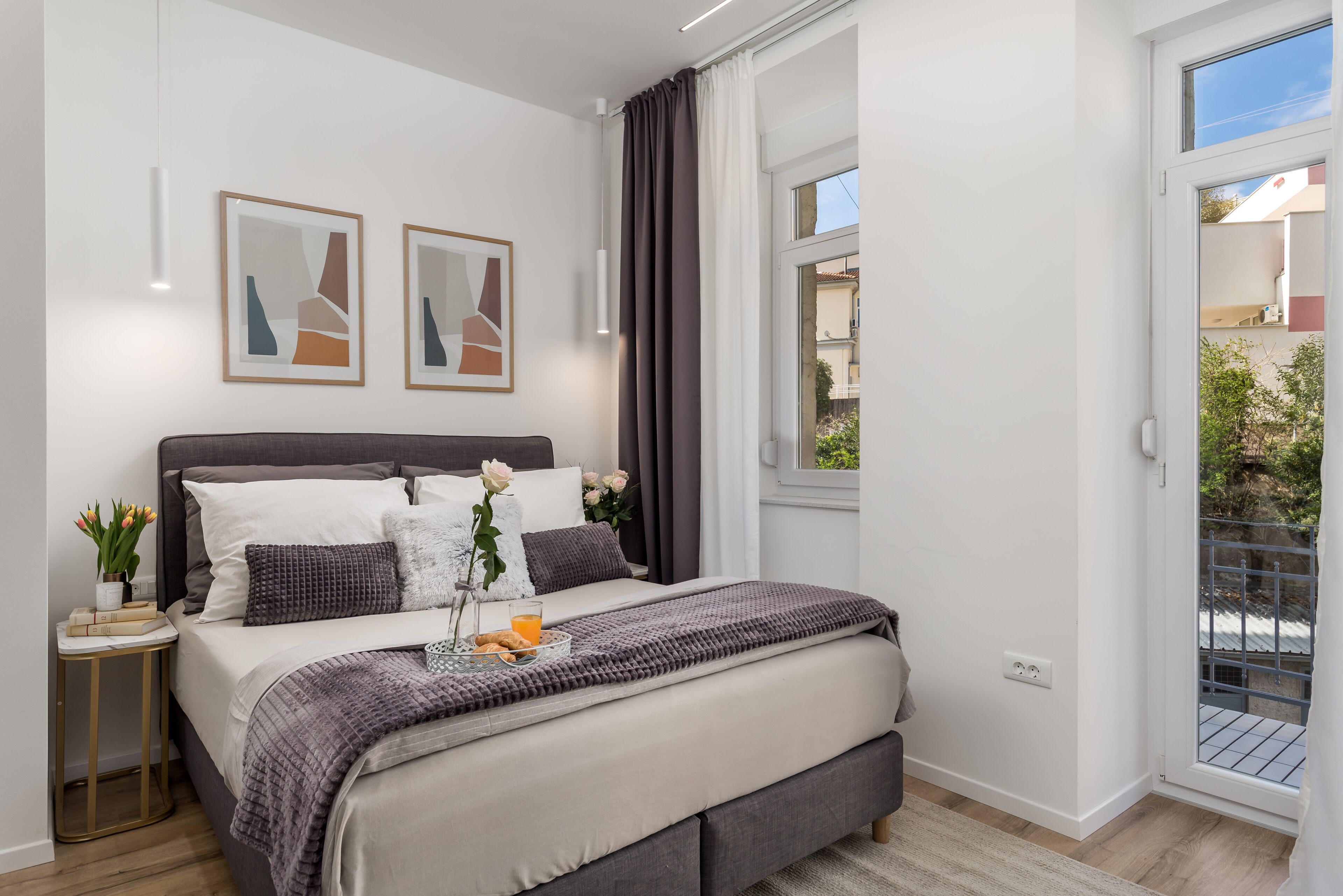 Ein modernes Schlafzimmer mit einem großen Bett, einem Nachttisch mit Büchern und Blumen sowie einem Fenster mit Vorhängen.