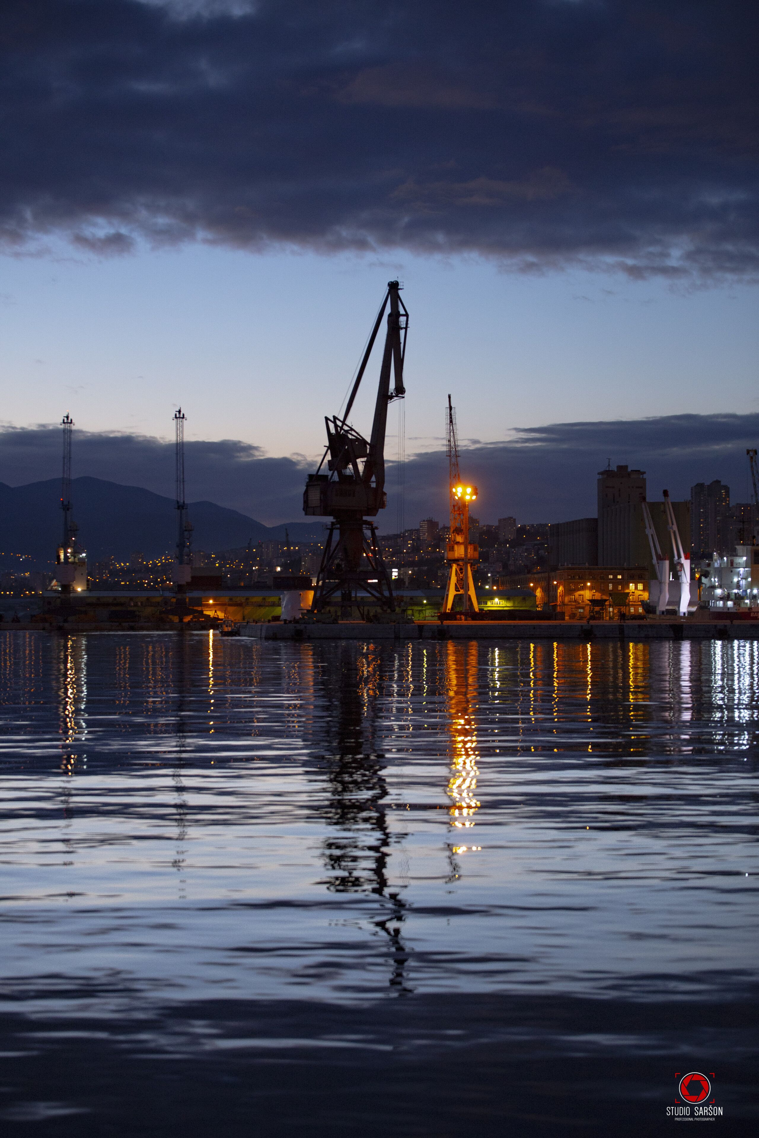 A waterfront cityscape at dusk with industrial cranes and buildings reflected in calm water.