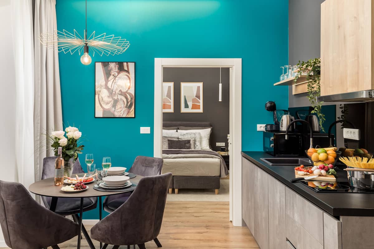 Une cuisine moderne avec un mur d’accent de couleur turquoise, un coin repas avec une table dressée pour deux personnes, et une chambre à coucher visible à travers une porte ouverte.