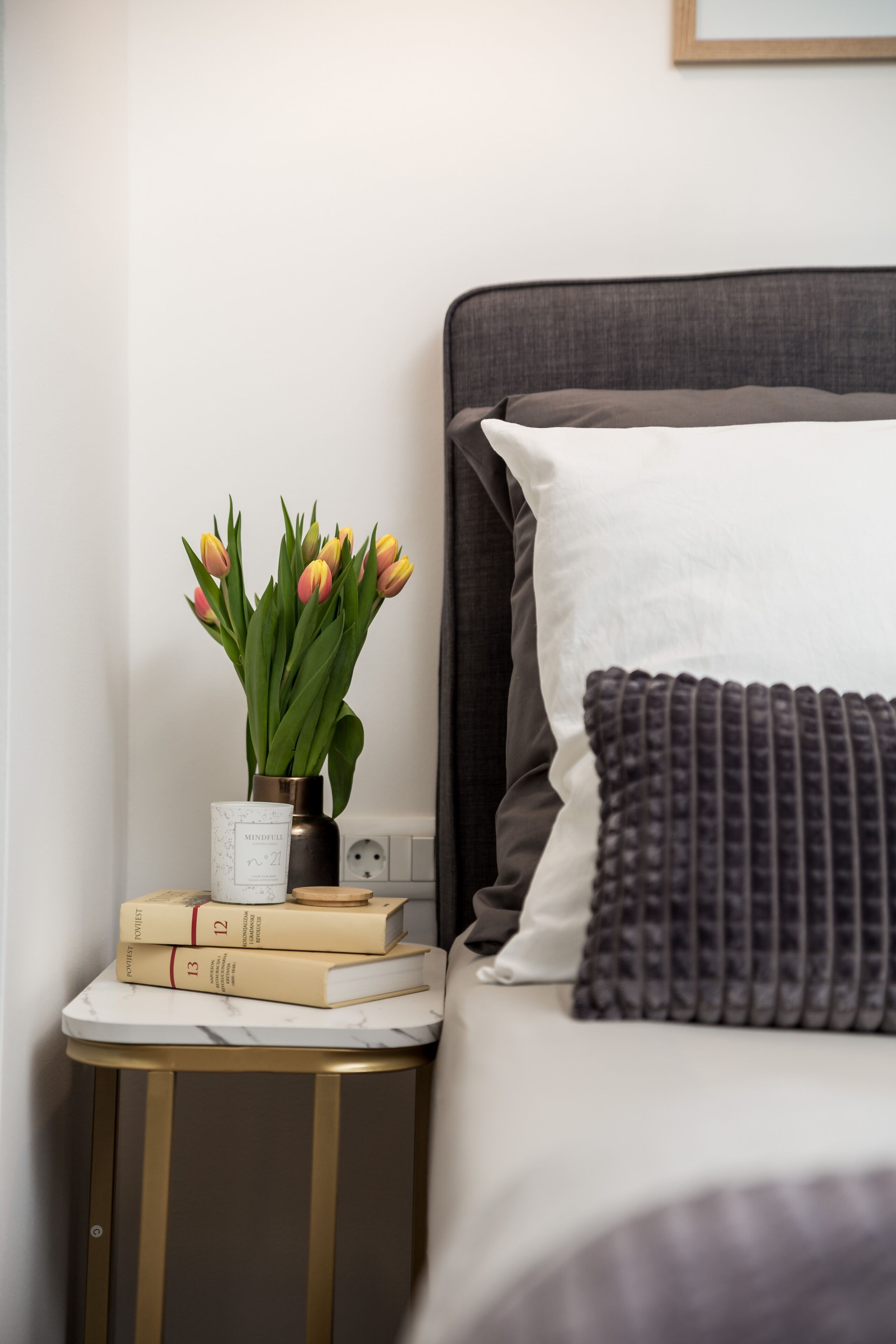 Una cama bien hecha, con cabecera gris y almohada blanca, una mesita de noche con un florero de tulipanes, y una pila de libros.