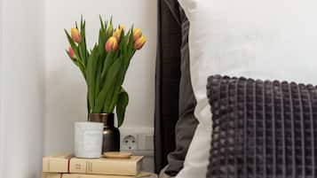 Una cama bien hecha, con cabecera gris y almohada blanca, una mesita de noche con un florero de tulipanes, y una pila de libros.