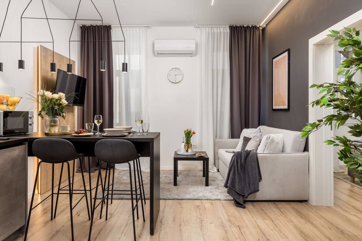 Un salon moderne comprenant un canapé, une table à manger avec des chaises, une petite table d’appoint avec un vase et une grande fenêtre avec des rideaux.