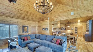 A cozy log cabin interior with a large blue sectional sofa, a chandelier, and a kitchen area. - Wee-Kin-Da-Wa | Getaway at Boyne Mountain | Skybridge View | Ski-In/Ski-Out (Boyne Falls)