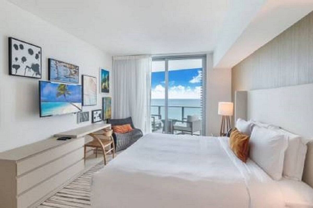 A hotel room with a large bed, a desk with a chair, a TV, and a balcony with a view of the ocean.