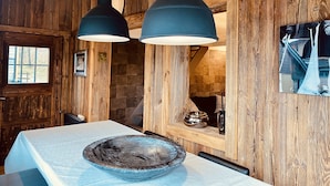 Un intérieur rustique avec des éléments en bois, une grande table, deux lampes suspendues et un mur en pierre.