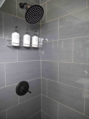 Ein Duschbereich mit Handbrause, am Wand montiertem Wasserhahn und drei Pflegeprodukten auf einem Regal.