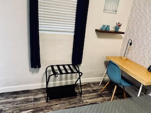 Ein Zimmer mit einem Holztisch, einem blauen Stuhl, einem schwarzen Metallregal und einem Fenster mit Jalousien.