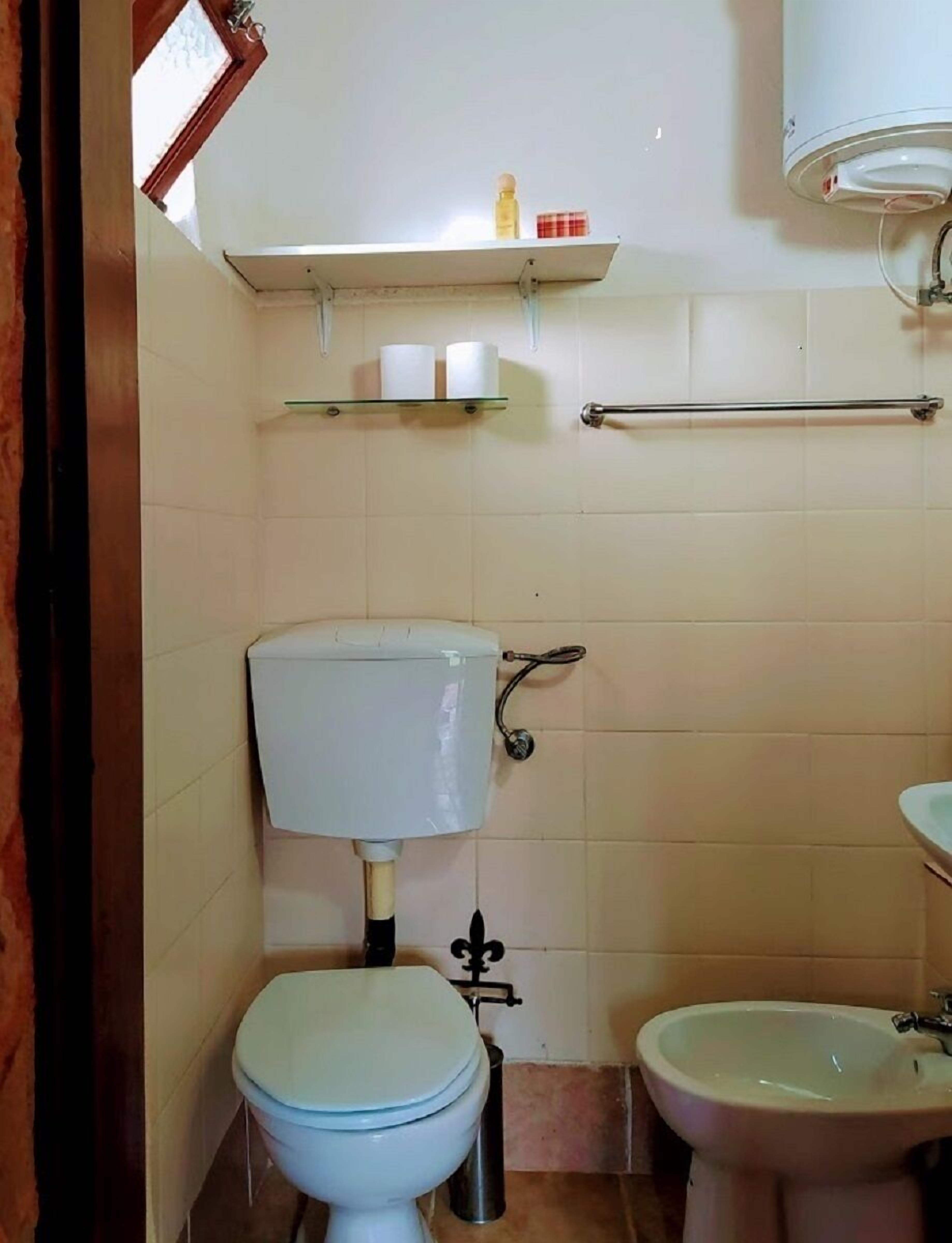 A small bathroom with a toilet, bidet, and sink. There is a shelf above the toilet with a bottle and a roll of toilet paper.