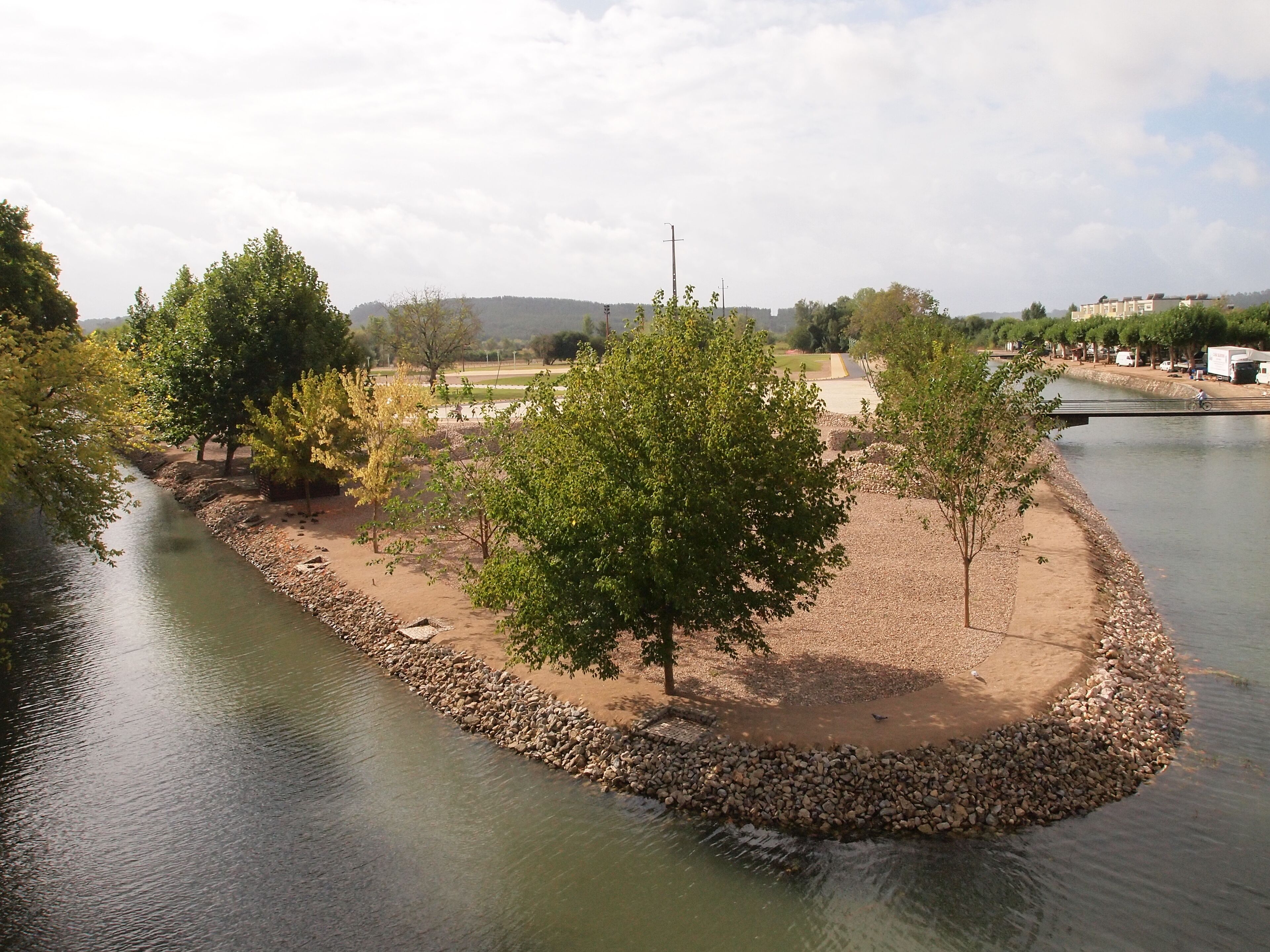 A riverside park with trees, a walking path, and a bridge.