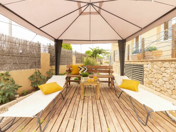 An outdoor patio with a wooden deck, white lounge chairs, a wooden table, and a large umbrella.