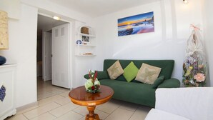 A living room with a green sofa, a wooden table with a vase of flowers, a white armchair, a white cabinet, and a wall-mounted shelf with decorative items. - Lovely apartment for 4 people with WIFI, pool, A/C, TV and terrace (Benissa)